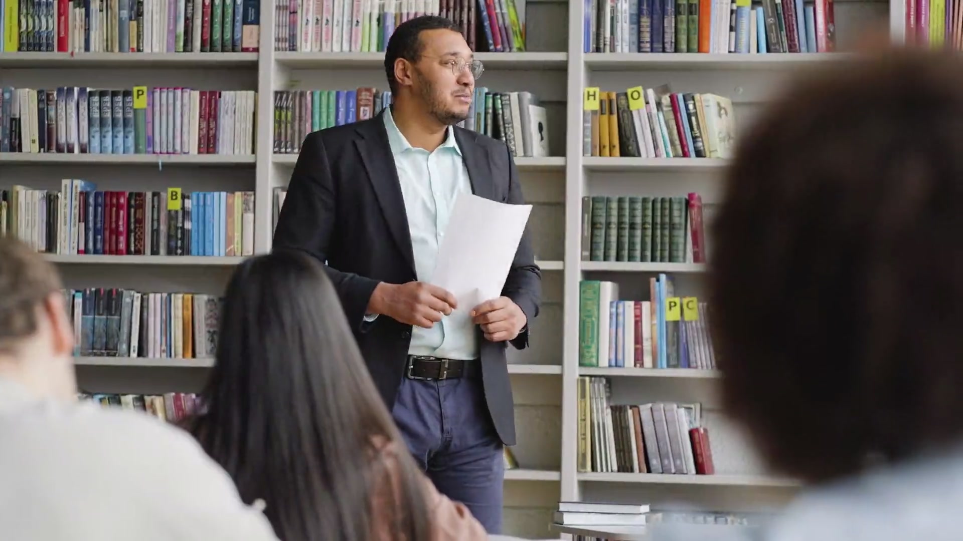 A teacher explaining a point to his class in the library, students listening in the foreground.